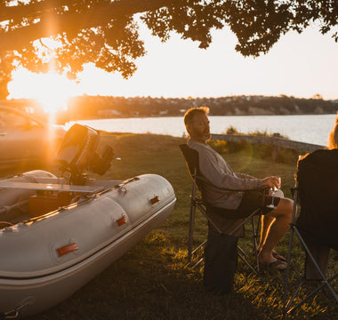 two people sitting in lawn chairs by the water with an inflatable boat
