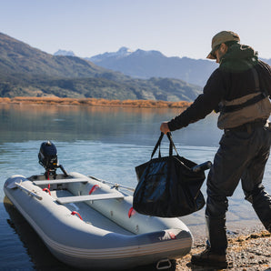 A person carrying a black bag towards a pearl grey inflatable fishing kayak with a small motor attached, parked on a beach with mountains in the background.